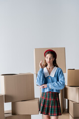 Stylish model in beret and knitted cardigan posing near carton boxes isolated on grey