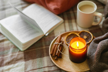 Burning candle with wooden wich in amber glass gar, open book and cup of coffee or tea. Autumn home decor. Cozy fall mood.