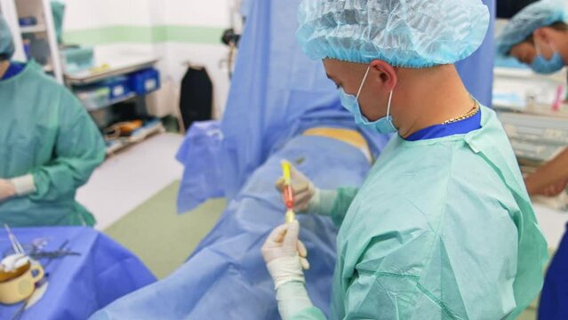 Male doctor holds a pair of syringes connected with each other. Specialist moves stem cells from one syringe to another. Patient prepared for operation lies on the operational table.