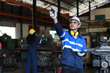 Technician engineer man in protective suit stand and use walkie talkie radio and tablet while controll or maintenance operation work lathe metal machine at heavy industry manufacturing factory