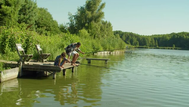 Joyful adorable school age multiracial boy and positive black father casting fishing rods from wooden pier on pond, talking and enjoying fishing together on summer day.