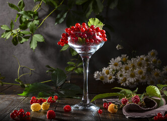 Fototapeta na wymiar Red currants in a glass wine glass, raspberries and a bouquet of daisies on a wooden table