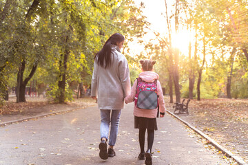 Mom and daughter walk in autumn park. Little girl after school with mother from back. Everyday life