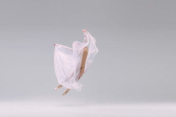 Naklejka premium Portrait of young ballerina dancing, performing in beige cloth, covered with fabric isolated over grey studio background