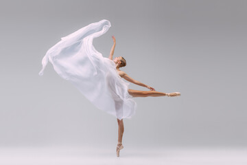 Portrait of young ballerina dancing with fabric isolated over grey studio background. Standing on tiptoe and throwing transparent cloth