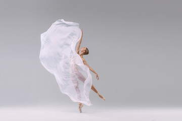 Portrait of young ballerina dancing with fabric isolated over grey studio background. Standing on tiptoe. Weightless transparent cloth