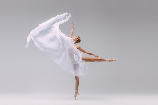 Fototapeta Portrait of young ballerina dancing with fabric isolated over grey studio background. Standing on tiptoe and throwing transparent cloth