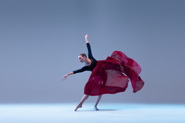 Portrait of young ballerina dancing with deep red fabric isolated over blue grey studio background. Artistic performance of classic dance