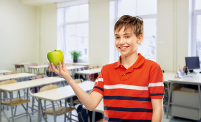 school, education and people concept - portrait of happy smiling student boy in red polo t-shirt holding green apple over empty classroom background