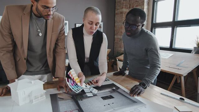Tracking out slow mo of three multiethnic female and male designers working on house exterior and interior design, standing around table in office at daytime, choosing paint color