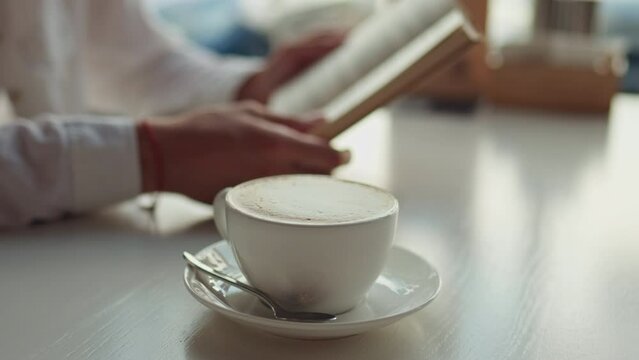 A cup of hot fresh cappuccino in a white dish stands on a white table against the background of a girl reading a book Girl reading book sitting on a summer bright day in a restaurant rest lunch snack
