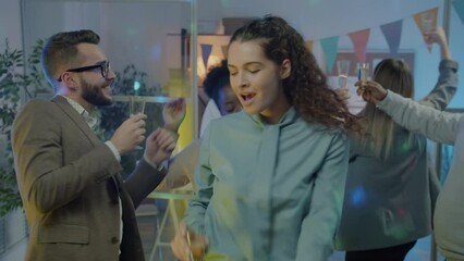 Happy young lady dancing with colleagues at corporate party holding glass of champagne in office at night. Youth and special occasion concept.