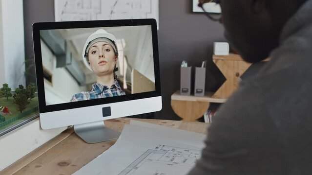 Over-shoulder of cropped architect standing at table in office at daytime, editing architectural plans, video calling female engineer at construction site