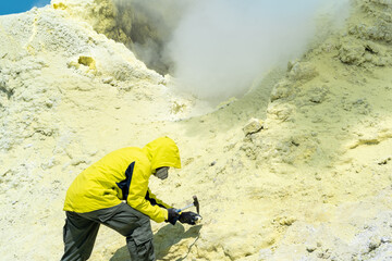 male volcanologist on the slope of a volcano next to a smoking sulfur fumarole examines a sample of...