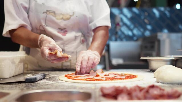 Chef prepares pizza spreads pieces of bacon on raw prepared dough, dressing preparing pizza for baking. Meat ingredients for pizza topping, Chef woman in white gloves prepares pizza
