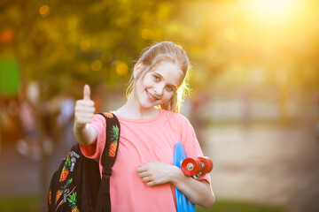 Smiling schoolgirl with backpack and skateboard showing thumbs up in sunlight. Educational course...