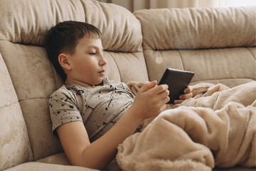 Teenage boy sitting on couch using tablet and playing video games on internet online at cozy home.