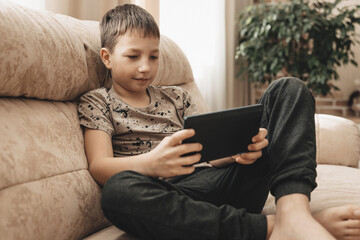 Teenage boy sitting on couch using tablet and playing video games on internet online at cozy home.