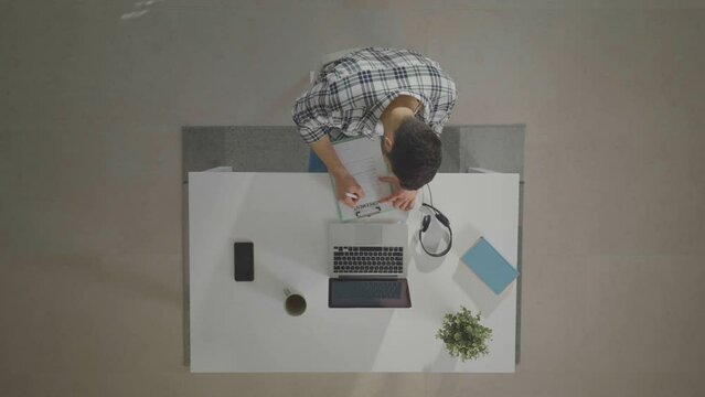 Caucasian man writing data into agreement. Top down view of light desk. Home office workroom from height.