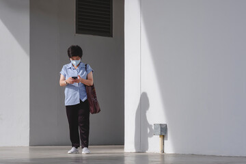 Asian woman in protective mask using smartphone while walking out of alley beside office building after work