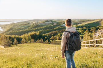 Naklejka na ściany i meble Tourist with backpack standing on top of hill in grass field and enjoying beautiful landscape view. Rear view of teenage boy hiker resting in nature. Active lifestyle. Concept of local travel