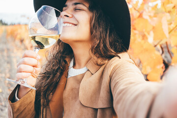 Smiling young woman in stylish hat and coat drinks delicious white wine from wineglass and making...