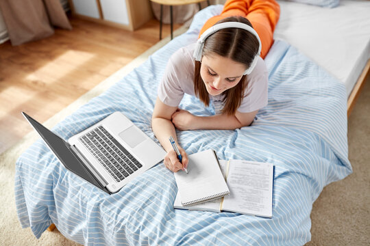 school, online education and e-learning concept - happy smiling teenage student girl in headphones with laptop computer writing to notebook lying on bed at home