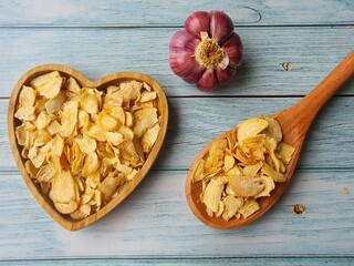 Thinly sliced fried garlic in a wooden heart-shaped bowl, a wooden spoon and a bulb of fresh garlic, isolated on a wooden table, top view, space for text.