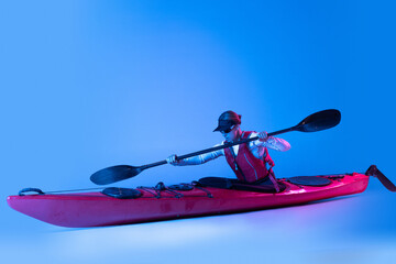 Canoeing. Sportive woman in red canoe, kayak with a life vest and a paddle isolated on blue background in neon light. Concept of sport
