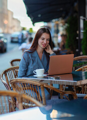 European business woman is working with laptop outdoors during coffee break, plans presentation, is focused on documents, looks through printed charts, wears business suit