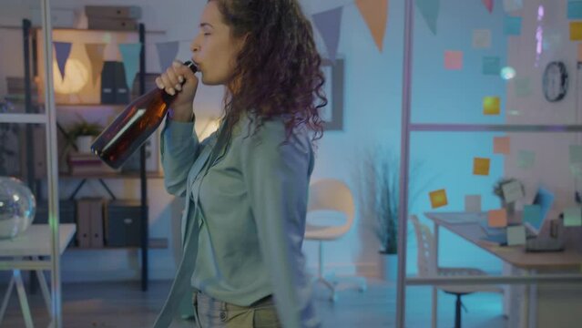 Slow motion portrait of happy young female entrepreneur dancing at work party and drinking champagne from bottle. Youth and holidays concept.