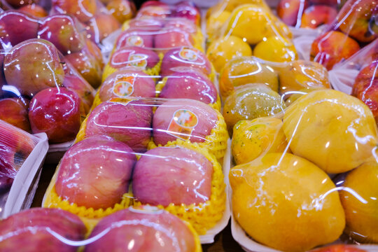 Assorted fresh fruits at market stall display wrapped in plastic film wrap for protection. The plastic wrappers are wasteful and unsustainable for the environment.