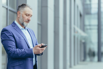 Serious and confident gray-haired man standing outside office building, businessman senior banker in business suit holding phone, successful investor using smartphone