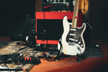 A white electric guitar and a red amplifier onstage with electrical audio cables and sound effects foot pedals - obrazy, fototapety, plakaty