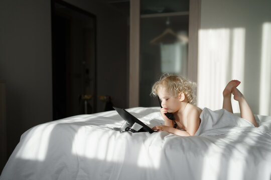 A blonde child with curly blond hair is talking on the phone in a hotel room lying on a bed in a towel. In the morning light