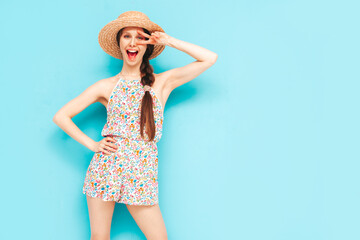 Portrait of young beautiful smiling female in trendy summer overalls. carefree woman posing near blue wall in studio. Positive model having fun indoors. Cheerful and happy. In hat. Shows peace sign