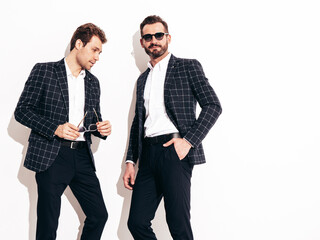 Portrait of two handsome confident stylish hipster lambersexual models. Sexy modern men dressed in black same elegant suit. Fashion male posing in studio near white wall. In sunglasses