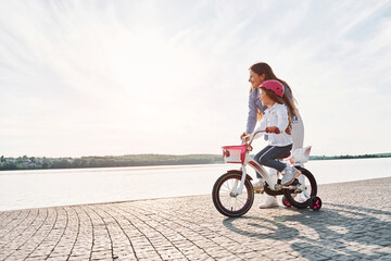 Side view. Near the lake. Mother with her young daughter is with bicycle outdoors together