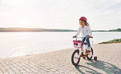 Fototapeta premium Near the lake. Happy little girl riding a bicycle outdoors in summer