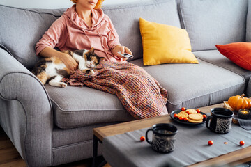 Woman with plaid selecting movie with remote controller and stroking multicolored cat pet on the...