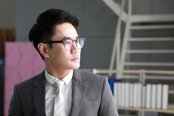 A young Asian businessman in a suit, wearing eyeglasses has the personality of a successful man standing in the office, looking outside.