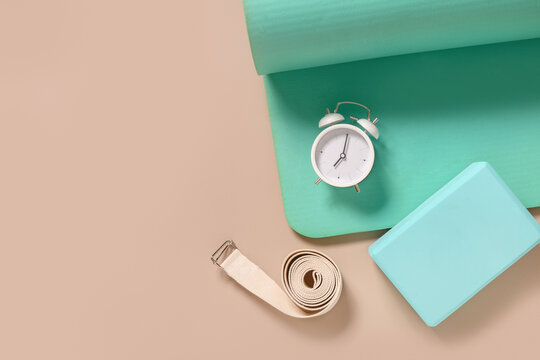 Green fitness mat, yoga blocks, clock and white belt on beige background. Yoga practice, relaxation and meditation concept. Top view with copy space.