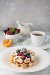 Waffles with berries and powdered sugar in a white plate in the background, fruits and a cup of tea...