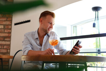 Successful rich businessman in shirt drinking cocktail and holding smartphone at table in restaurant