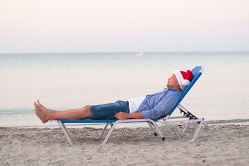 Christmas party, Xmas time, festive . Seasonal holidays. Single young man in Santa hat on chaise-longue on sand of the sea, soft focus, close up view, isolated on blue. Bottle of whiskey in Santa hat