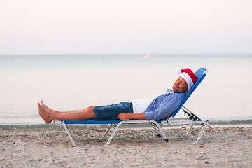 Single young man in Santa hat on chaise-longue on sand of the sea. Christmas party, Xmas time,...