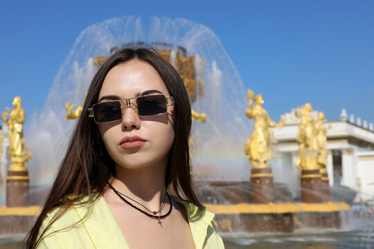 Attractive girl with long hair wearing sunglasses standing on summer fountain background. Female beauty, tourist in VDNKh park in Moscow in sunny day