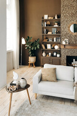 Contemporary beige sofa with coffee table on the backgroung of brown wall with fireplace, bookshelf with decor and potted plants in the living room with large windows. Cozy chalet interior.