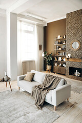 Modern stylish living room with large windows and sofa with pillow and plaid on the backgroung of brown wall with brick fireplace, shelving with books, decor and potted plants. Cozy chalet interior. 
