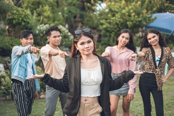 Four young people point to their confident and popular leader, who shrugs. A youthful and upbeat team of five posing outdoors.